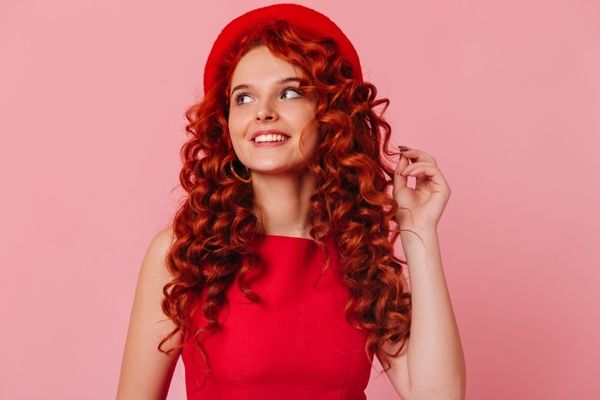 Affascinante ragazza dai capelli rossi con il sorriso distoglie lo sguardo. La donna con cappello e top rosso si tocca i capelli. negozi di parrucche a Roma