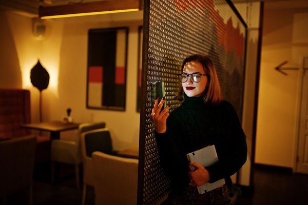 Sala di fuga misteriosa XR Allegra giovane bella donna rossa con gli occhiali che utilizza un telefono cellulare in una stanza buia con lo schermo del telefono visualizzato sul viso 