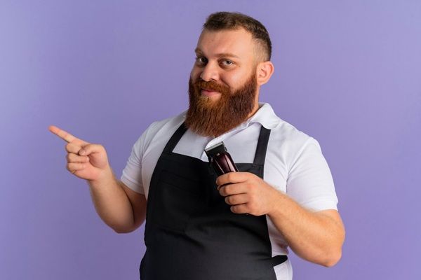 Barbiere professionista in grembiule con la macchina per tagliare i capelli che punta il dito verso il lato sorridente in piedi sul muro viola barbieri di Roma